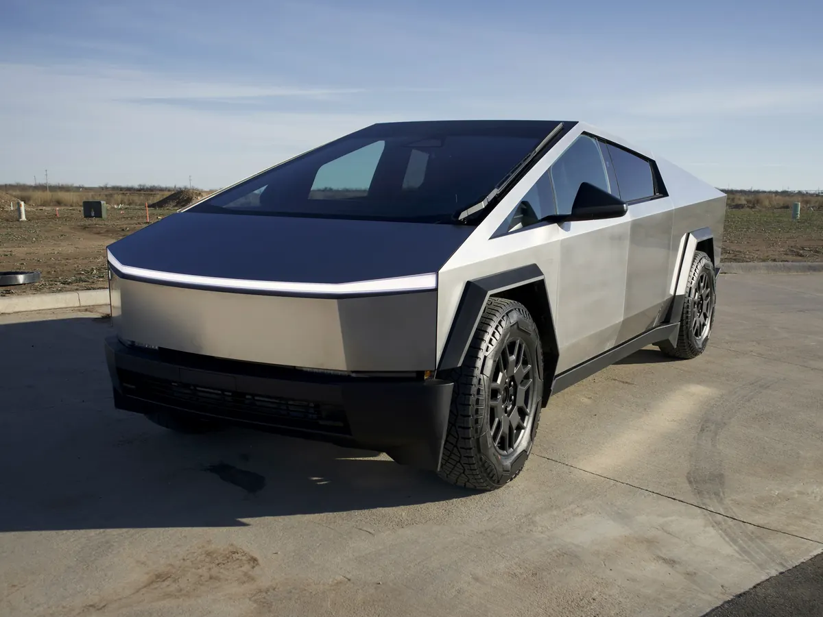 Stainless-steel Tesla Cybertruck in front three-quarter view, with its full-width light bar, in an open car park