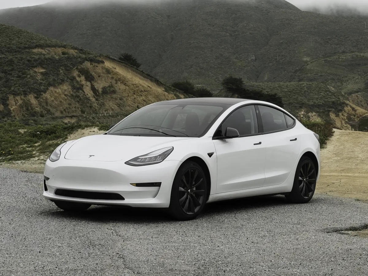 White Tesla Model 3 (2017–2023) in front three-quarter view on a gravel lay-by, with misty green hills behind