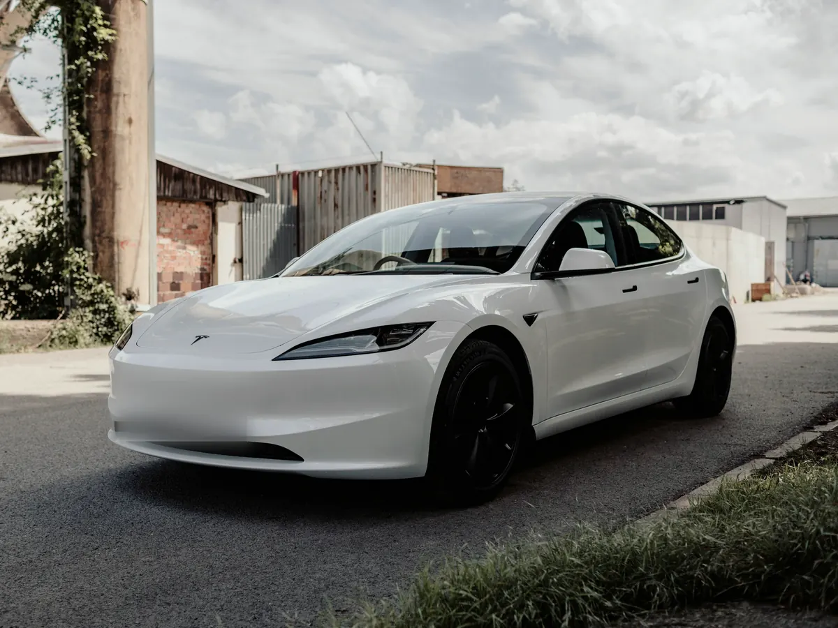 White Tesla Model 3 “Highland” (2024+) with slim headlights, front three-quarter view on a quiet industrial street