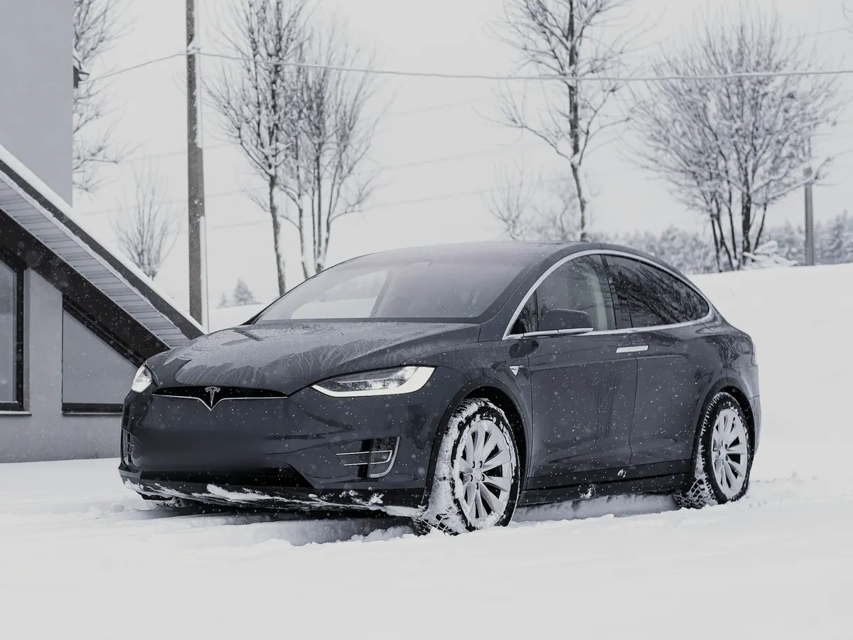 Dark grey Tesla Model X (2015–2020) in front three-quarter view, parked in fresh snow beside a modern building