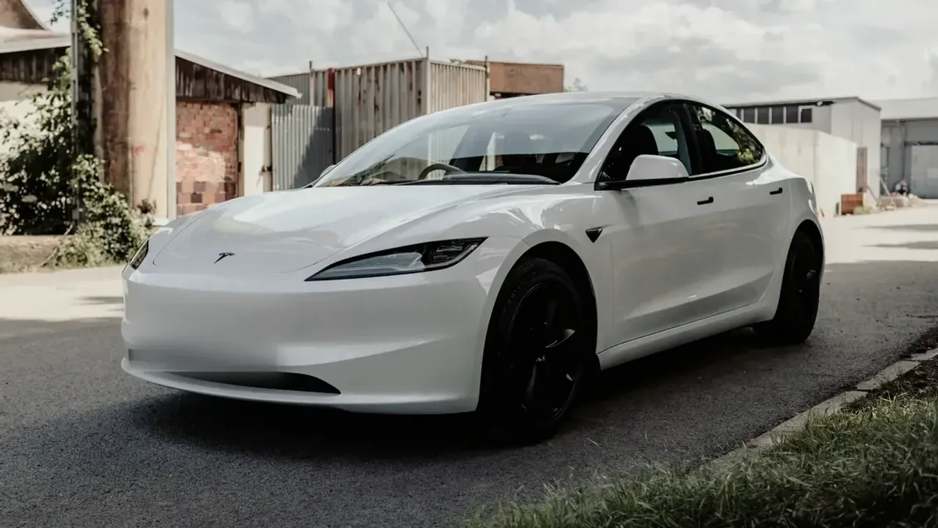 White Tesla Model 3 “Highland” (2024+) with slim headlights, front three-quarter view on a quiet industrial street