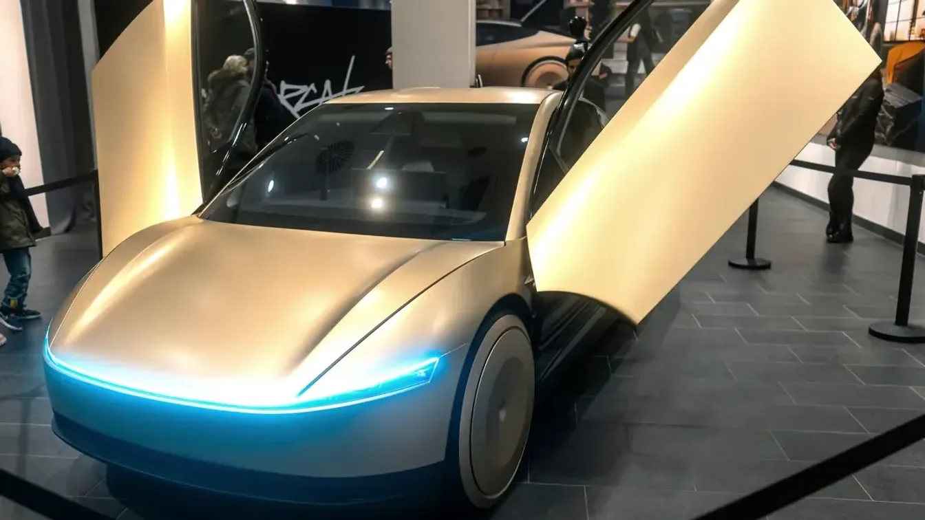 Gold Tesla Cybercab two-seat robotaxi in front three-quarter view, with both butterfly doors raised and its full-width front light bar glowing, on display in a Berlin Tesla store
